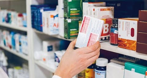Mano revisando una caja de medicamento frente a estanterías de farmacia