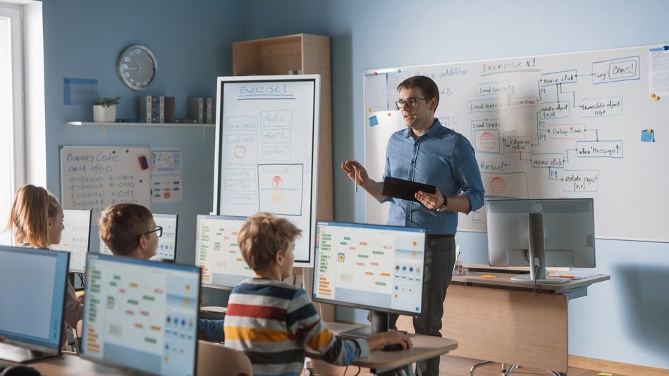 Profesor explicando diagramas en la pizarra a estudiantes frente a computadores