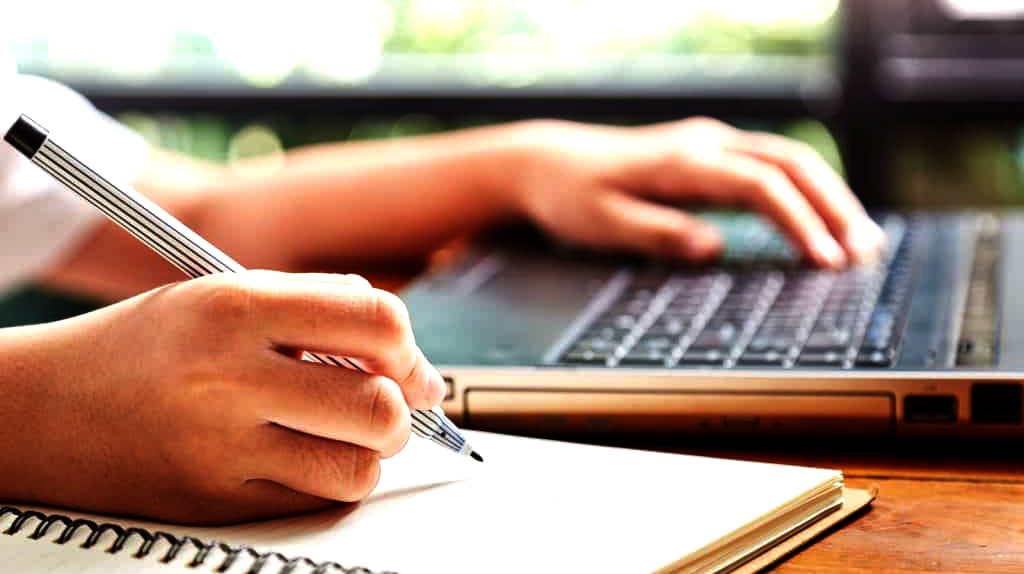 Manos escribiendo en una libreta junto a un computador portátil