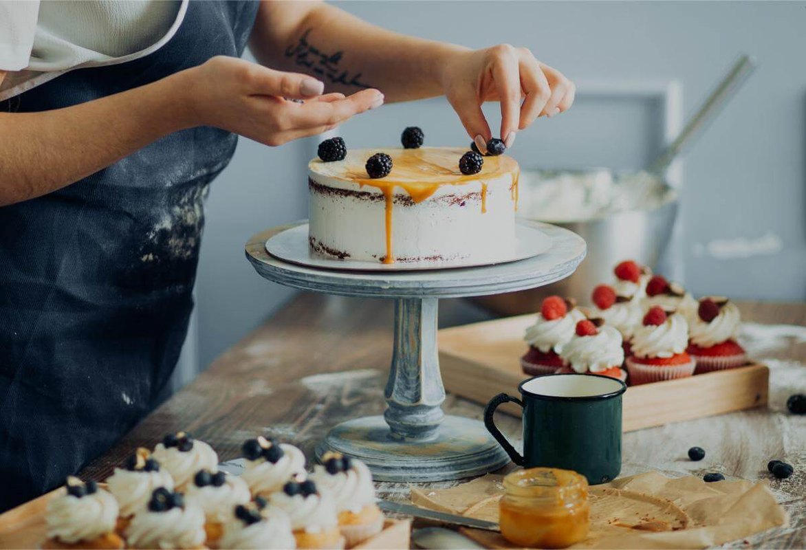 Repostera decorando con moras una torta con caramelo, rodeada de cupcakes