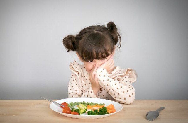 Niña pequeña tapándose la boca frente a un plato de verduras